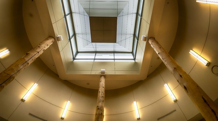 Looking up at a round rotunda with three large round logs holding up the structure with a large skylight above and long vertical lights around the circle.