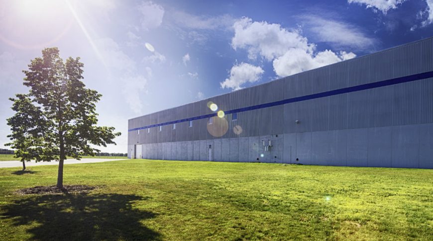 Exterior view of long precast concrete building with blue stripe on top third of building and seven windows on top half of building with one garage door to far end of building. Grassy area with a couple trees in foreground with sun-kissed blue sky with fluffy clouds in background.