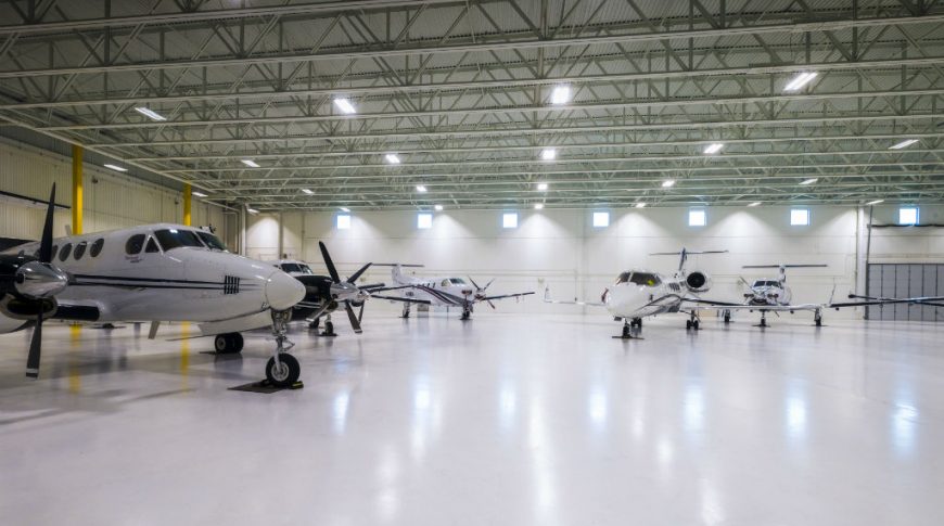 Interior of hangar building with light white floors and white metal ceiling overhead with fluorescent lights and private jets throughout.