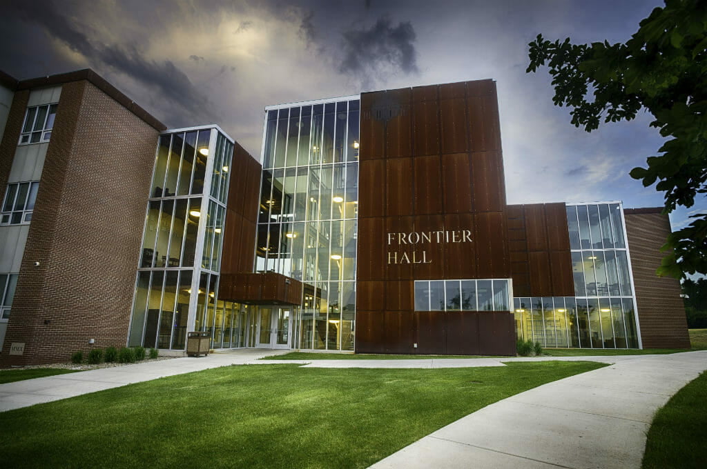 Close-up exterior view of the front of Frontier hall showcasing 4 levels of floor-to-ceiling windows fand brown paneling with the words "Frontier Hall" on the face of the paneling and brick building attached to the far left side.