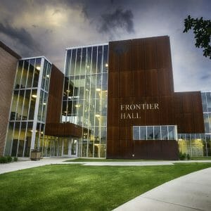 Close-up exterior view of the front of Frontier hall showcasing 4 levels of floor-to-ceiling windows fand brown paneling with the words "Frontier Hall" on the face of the paneling and brick building attached to the far left side.