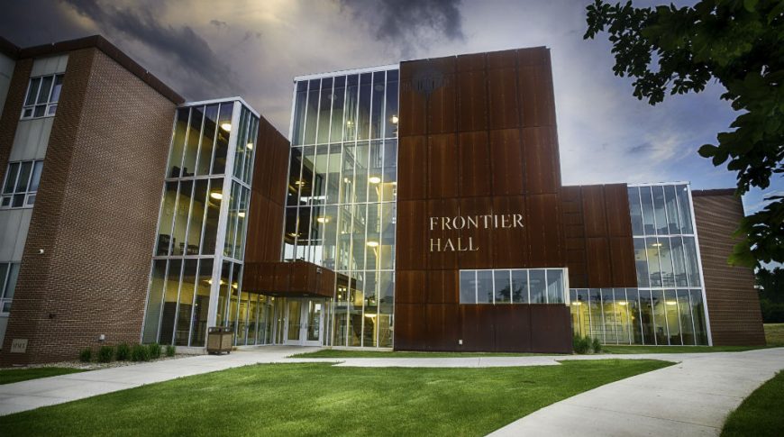 Close-up exterior view of the front of Frontier hall showcasing 4 levels of floor-to-ceiling windows fand brown paneling with the words "Frontier Hall" on the face of the paneling and brick building attached to the far left side.