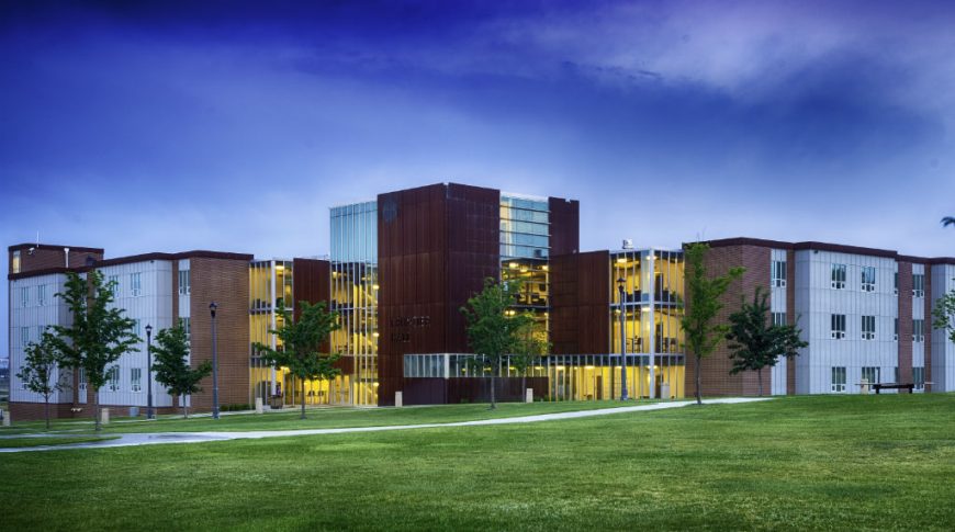 Far back view of Frontier Hall from across the grassy knoll at dusk with warm yellow lights shinning behind floor-to-ceiling walls on the modern building.