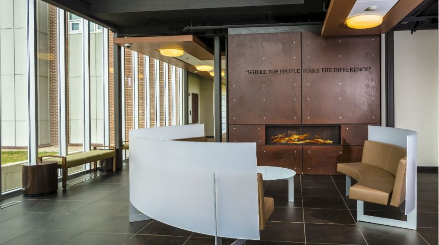 Modern room with wall of floor-to-ceiling windows to the back right a rusted metal clad fireplace to the back right and futuristic semi-circle couches and table to the foreground.