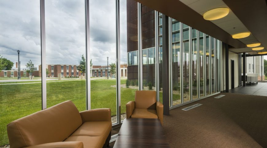 Interior of building facing wall of floor-to-ceiling windows with light brown leather coach and chair with brown table in foreground and hallway to back left. Outside in the background is green grass and brick buildings in the background.