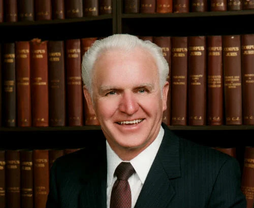 LeMar_Lynn-500x410 Head and shoulders portrait of owner Lynn Comstock who is an older white male with white hair wearing black suite, white-collared shirt and dark maroon and polka dot tie with library book backdrop