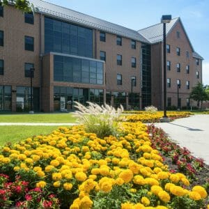 Exterior view of four-story brown brick learning-living college center with grassy knoll in front and concrete pathways bordered with bright yellow marigold and other flowers.