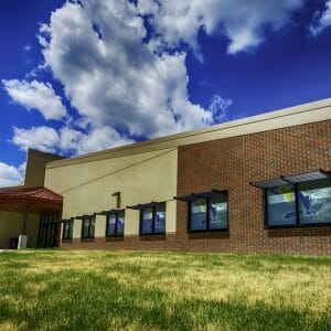 Exterior corner view from a low-height angle of the brick and beige concrete school building with grassy knoll in foreground and deep blue sky with fluffy clouds in background and school eagle mascot decorating each window.