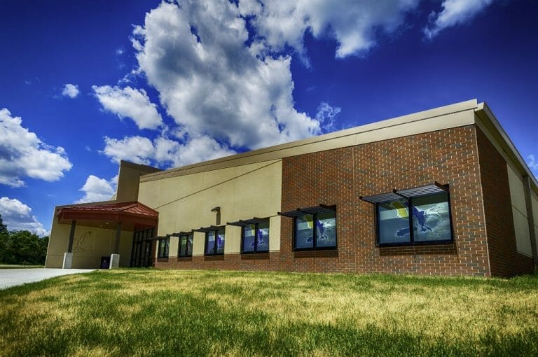 Exterior corner view from a low-height angle of the brick and beige concrete school building with grassy knoll in foreground and deep blue sky with fluffy clouds in background and school eagle mascot decorating each window.