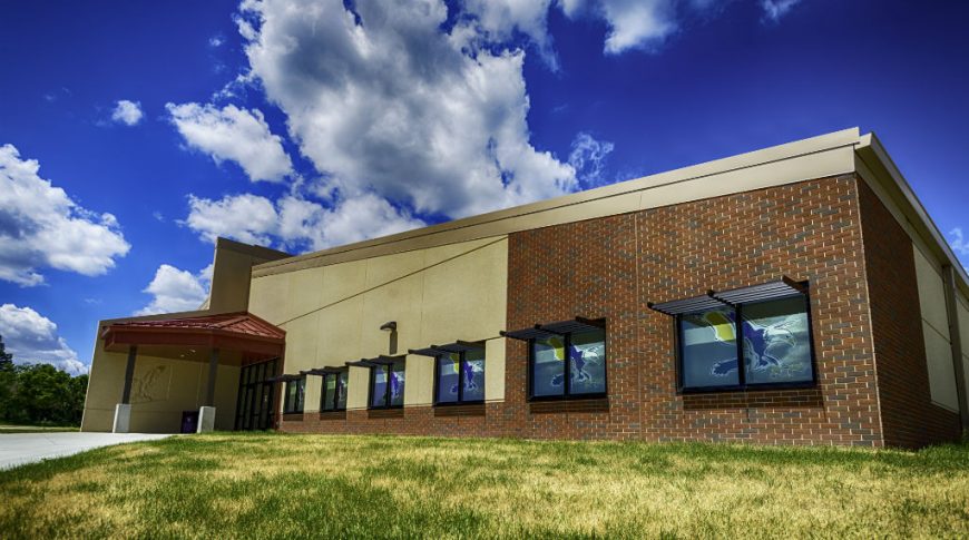 Exterior corner view from a low-height angle of the brick and beige concrete school building with grassy knoll in foreground and deep blue sky with fluffy clouds in background and school eagle mascot decorating each window.