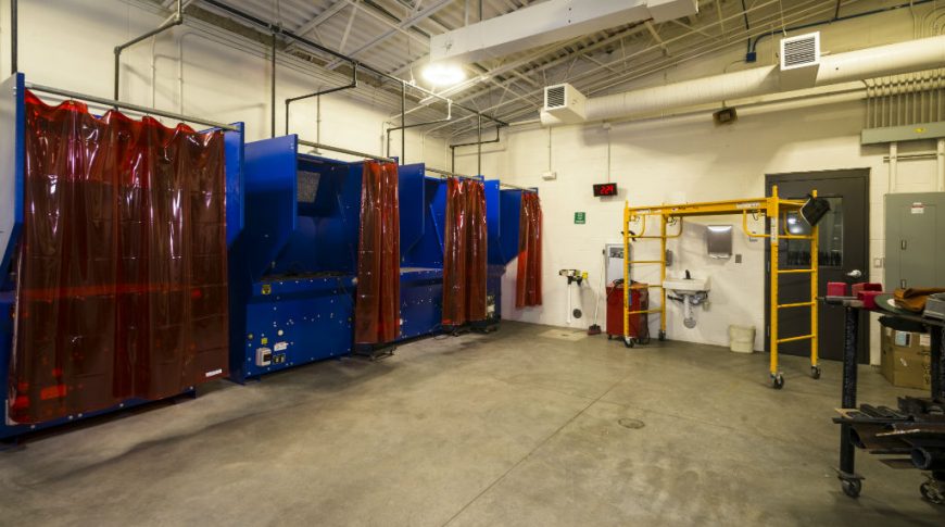 Beige-painted cinder block room with concrete floors and big blue machinery and red plastic curtains on the back left wall with door, electric panel and yellow scaffolding on back right wall.