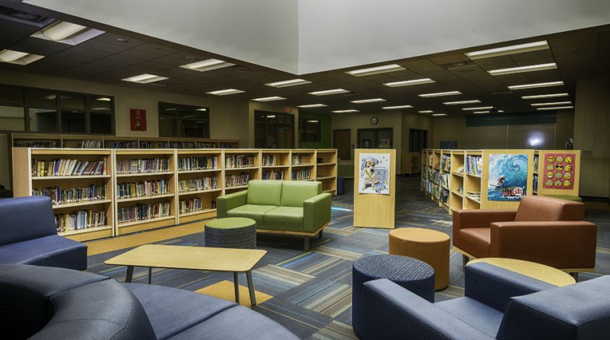 Back corner of library lounge area with dark navy, green, and brown chairs with light maple bookshelves behind and large skylight above.
