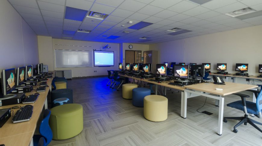 Long room with long light birch tables going down the far left and right sides and center with computer workstations down each one and chairs underneath each station. Projector and whiteboard to the far back wall.