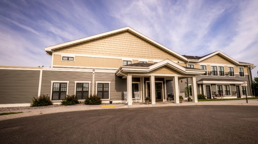 Front exterior view of two-toned beige upper vertical siding and cape-code shingling with taupe lower horizontal siding. Entrance covered porch with four columns leading with dark gray asphalt parking lot in foreground.