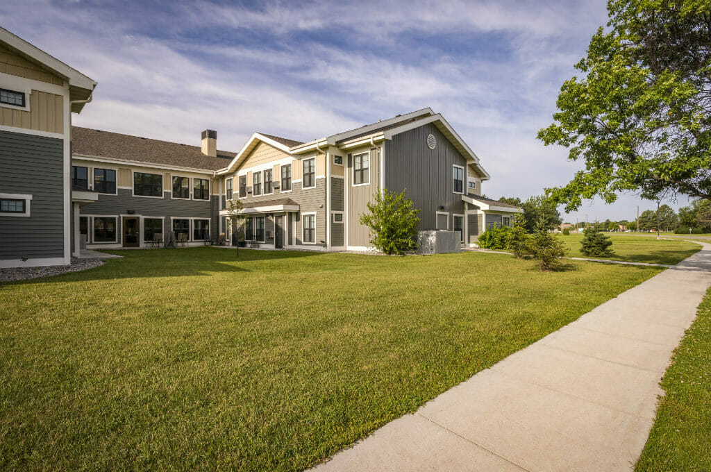 Exterior view of Siena Court Assisted Living building with two-tone grey/green and beige siding with lush green lawn and trees throughout.