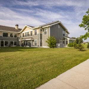 Exterior view of Siena Court Assisted Living building with two-tone grey/green and beige siding with lush green lawn and trees throughout.