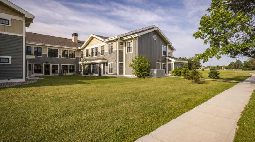 Exterior view of Siena Court Assisted Living building with two-tone grey/green and beige siding with lush green lawn and trees throughout.