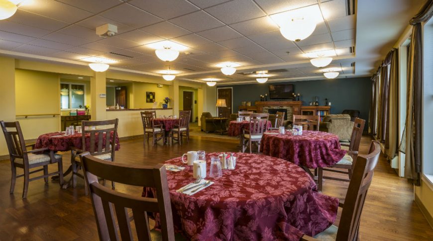 Dining room with five small round tables with burgundy leaf tablecloths and two wooden chairs at each with a fireplace and reading nook to the back wall.