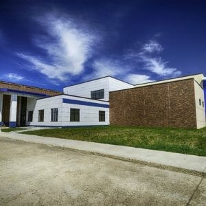 Exterior view of school from parking lot shows angular building with precast concrete panels, brick walls, and blue siding mixed. grassy area in foreground and deep blue sky to the background.
