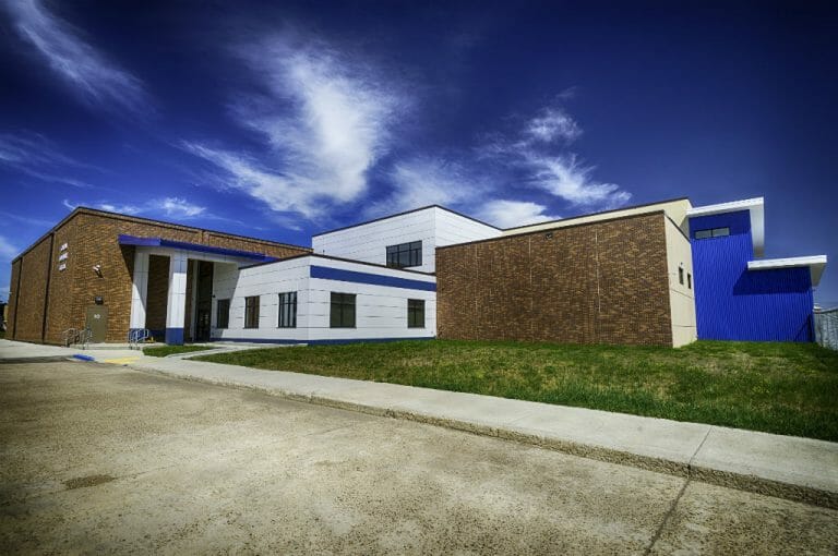 Exterior view of school from parking lot shows angular building with precast concrete panels, brick walls, and blue siding mixed. grassy area in foreground and deep blue sky to the background.