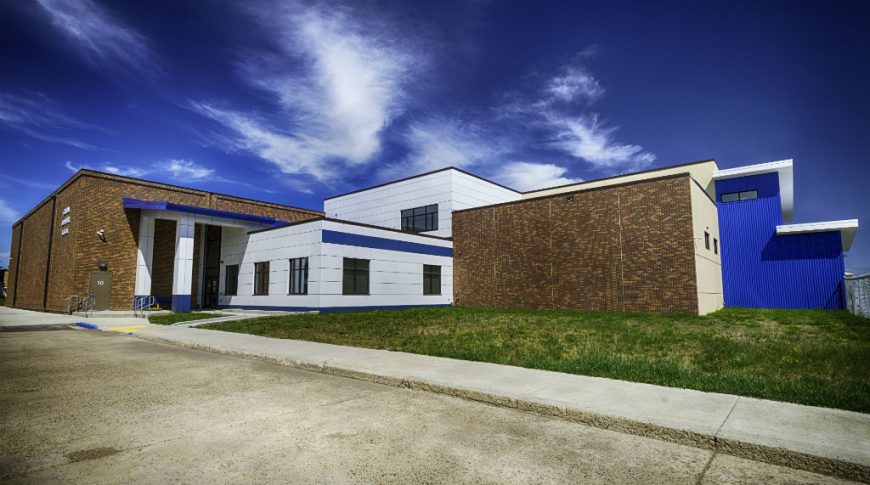 Tioga High School-P-Ext1-1024x680-Education Construction Exterior view of school from parking lot shows angular building with precast concrete panels, brick walls, and blue siding mixed. grassy area in foreground and deep blue sky to the background.