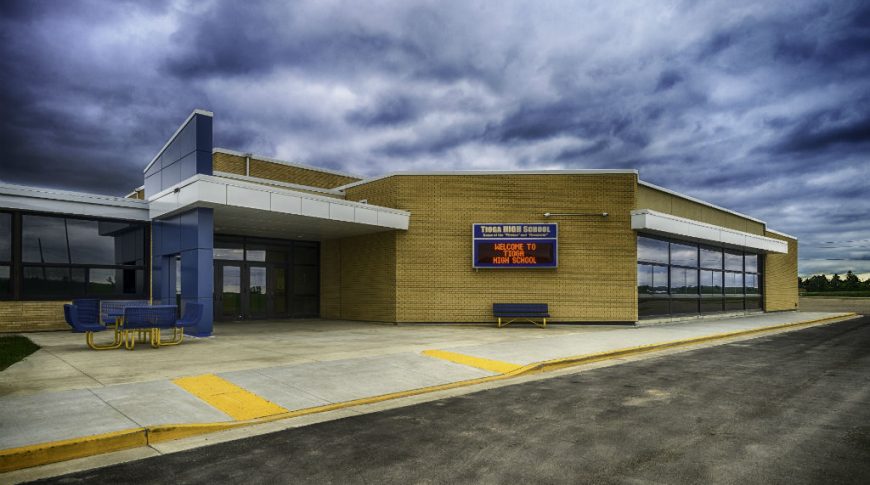 Tioga High School-P-Ext3-1024x680-Education Construction Exterior of yellow bricked high school with blue and silver entryway arches, blue and yellow metal picnic table and bench and digital sign board on the outside of the building.