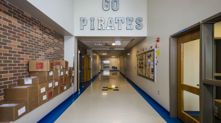 Tioga High School-P-Int1-1024x680-Education Construction Long hallway with beige walls and metal letters saying "Go Pirates" overhead with brick wall to the left side.