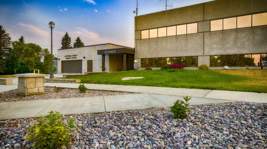 Exterior view of the Bottineau County Jail entrance. Features a precast concrete exterior with green lawn in front of the entrance and rock and sidewalk landscaping to the foreground