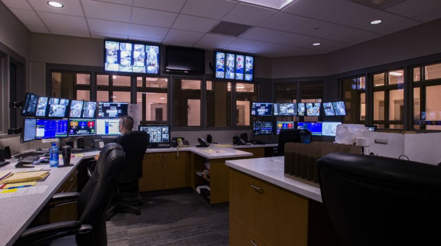 Jail surveillance room with glass windows on each side and computer stations with many security camera views to the far back.