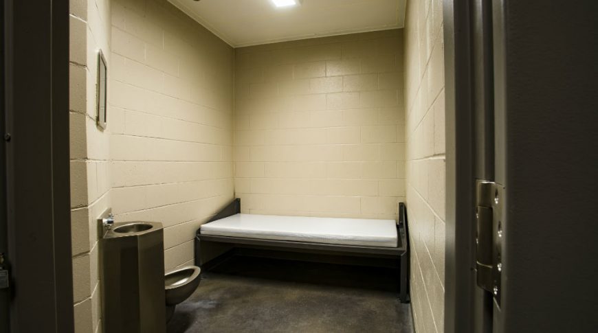 Beige-colored cinder block jail room with bed and mattress on the back wall and sink and toilet to the left wall.