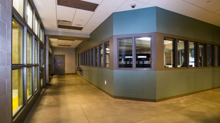 Jail hallway with concrete floors, paneled ceiling tiles, and drinking fountains to the end of the hallway. In the middle is a glass windowed section with teal-colored wall accenting.