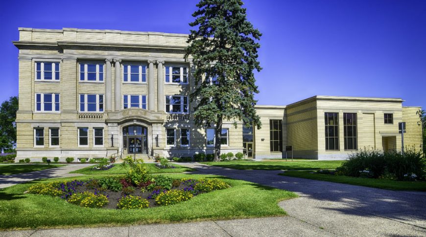 OtterTail Courthouse-P-Ext1-1024x680-Government Construction Exterior of beige brick 3-story courthouse with tall pine tree towering overhead and grassy knoll with flowers and sidewalks to the front