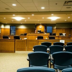 Courtroom with blue chairs for viewers, wooden cabinetry throughout the front with judge's podium and flags and windows on either side