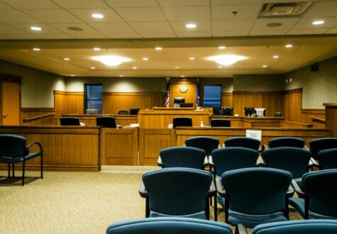 Courtroom with blue chairs for viewers, wooden cabinetry throughout the front with judge's podium and flags and windows on either side