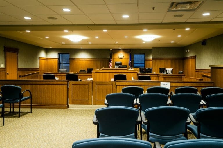 Courtroom with blue chairs for viewers, wooden cabinetry throughout the front with judge's podium and flags and windows on either side