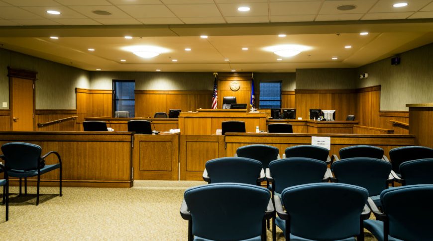 OtterTail Courthouse-P-Int1-1024x680-Government Construction Courtroom with blue chairs for viewers, wooden cabinetry throughout the front with judge's podium and flags and windows on either side