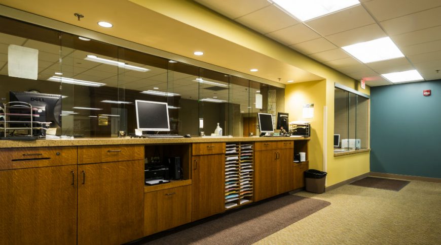 OtterTail Courthouse-P-Int3-1024x680-Government Construction Behind-the-glass view of courthouse support desks with brown cabinetry, speckled brown countertops, brown carpeting and yellow and blue accent walls