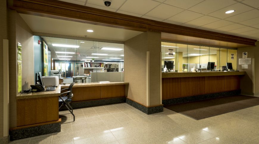 OtterTail Courthouse-P-Int4-1024x680-Government Construction Front reception desks with brown paneling, speckled brown countertops and counter-to-ceiling glass barrier with glimpses of private office areas behind the glass.