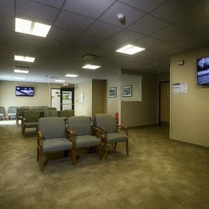 Beige-speckled wall waiting room with beige carpet tiles and chairs throughout with a couple of big-screen TVs on the far right and back walls