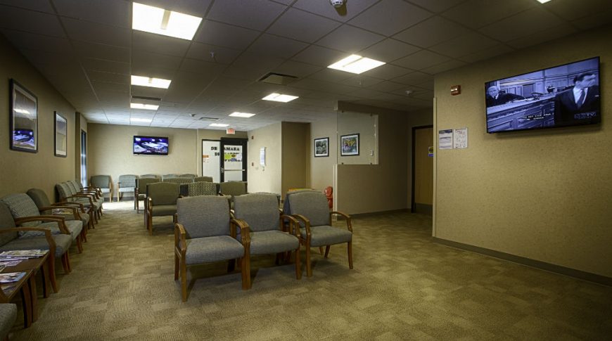 Beige-speckled wall waiting room with beige carpet tiles and chairs throughout with a couple of big-screen TVs on the far right and back walls