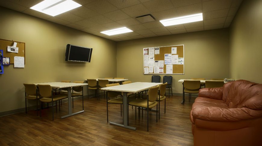Light brown walled room with fluorescent lighting and paneling above with dark brown faux-wood floors. Tables and chairs throughout with a brown leather cough to the right side