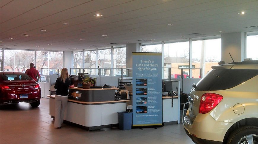 Inside vehicle dealership showing service desk in the middle of the room with office cubicles behind and two vehicles to each side
