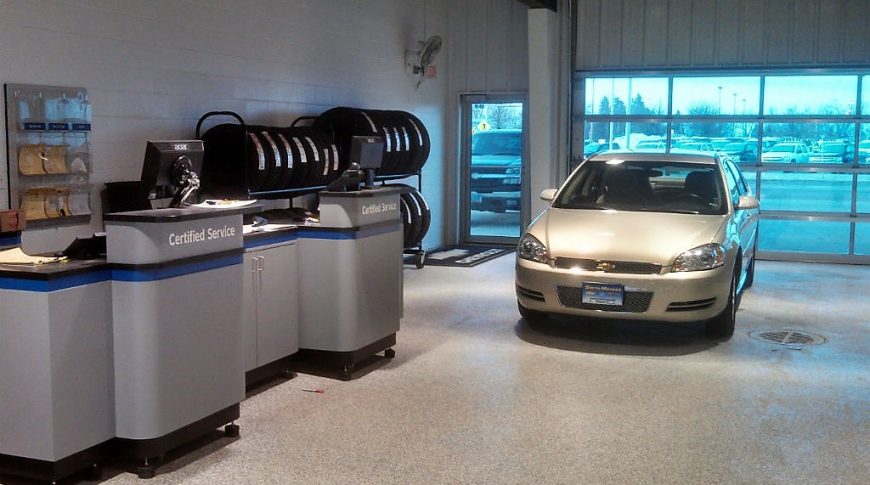 Inside glass garage door of vehicle service center with gray and blue-accented cabinetry to the left wall with tires behind and a car waiting to be serviced