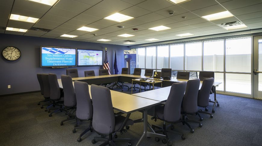 Southwest Water-P-Int1-1024x680-Government Construction Conference room with dark gray floor squares, square open table to the center of the room with dark gray office swivel chairs around, two large tv monitors to the back wall and wall of windows on the right wall