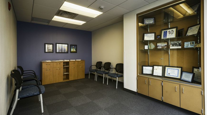 Southwest Water-P-Int2-1024x680-Government Construction Waiting area with dark gray carpeted floor tiles with wood paneled and glass showcase cabinet built in to beige wall to the left and dark blue accent wall to the back with chairs throughout