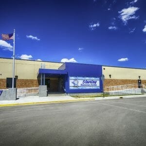 Exterior parking lot view of brown and beige concrete school entrance with bright blue bump-out entrance and flagpole to the left