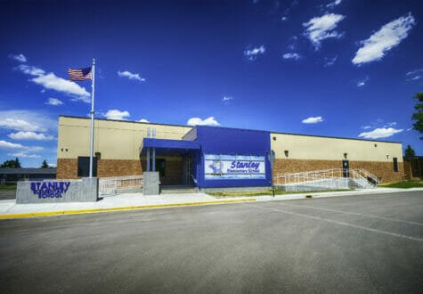 Exterior parking lot view of brown and beige concrete school entrance with bright blue bump-out entrance and flagpole to the left