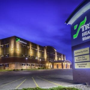 Exterior view of Trinity Health Center in Williston, ND showcasing lit up brown brick façade at dusk with lit up sign to the right in the foreground