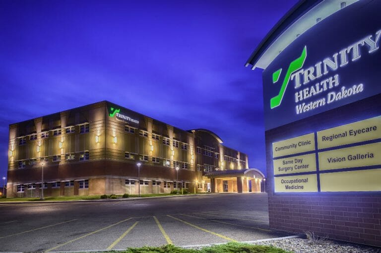Exterior view of Trinity Health Center in Williston, ND showcasing lit up brown brick façade at dusk with lit up sign to the right in the foreground