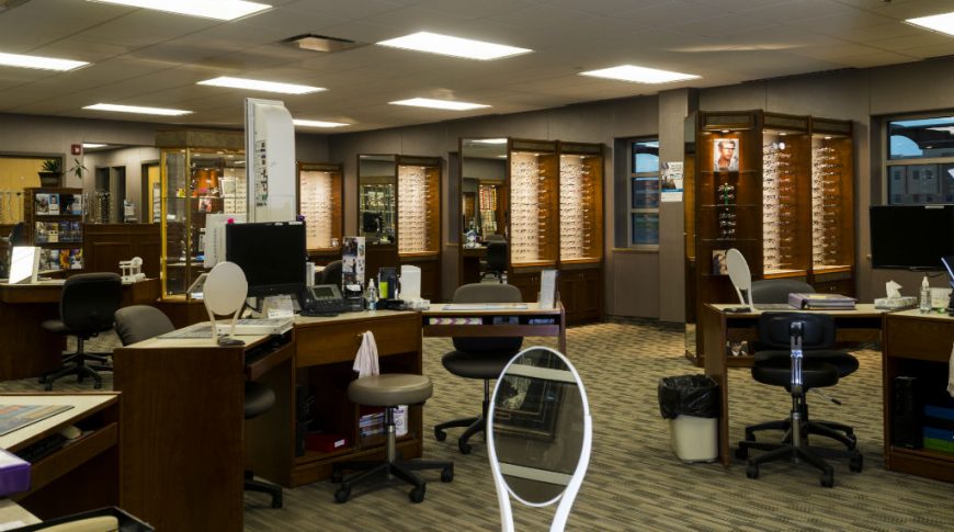 Trinity Health Western Dakota-P-Int1-1024x680-Healthcare Construction Vision center with glasses frames displayed in dark wooden cabinetry to the back of the room with desk stations to the middle of the room